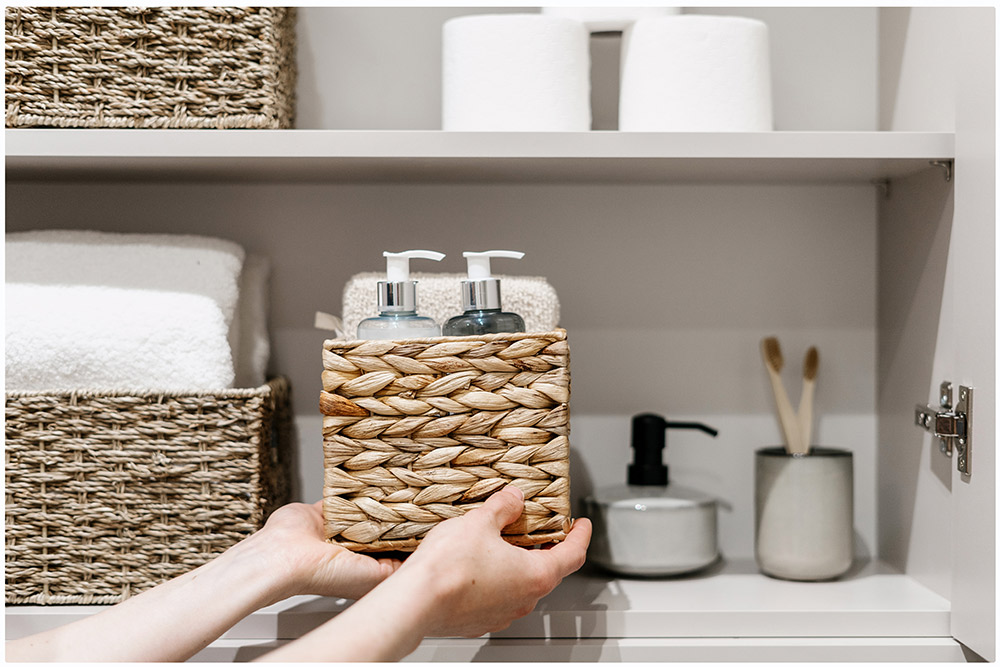 The image shows a shelf of the bathroom closet with all the things organized in different baskets of natural fibers and other containers. The colors of the closet and containers are neutral: gray, white and brown.